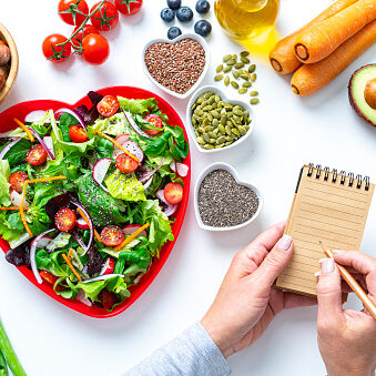 Overhead view of a red heart shape plate with healthy salad shot on white background. Water bottle, dumbbells and a tape measure are at the top right. Female hands are writing with a pencil on a note pad. Multi colored fresh fruits, vegetables, seeds and nuts are around the salad plate. High resolution 42Mp studio digital capture taken with SONY A7rII and Zeiss Batis 40mm F2.0 CF lens